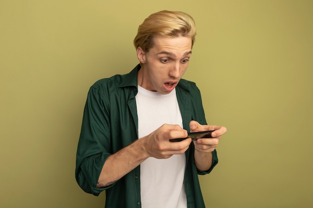 Verraste jonge blonde kerel die groen t-shirt draagt dat op telefoon speelt