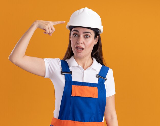 Verraste jonge bouwersvrouw in uniforme punten op zichzelf geïsoleerd op oranje muur