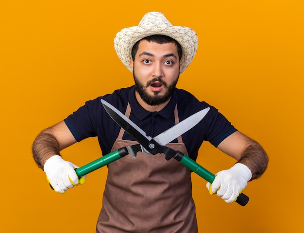 Gratis foto verraste jonge kaukasische mannelijke tuinman die een tuinhoed en handschoenen draagt die een tuinschaar vasthoudt en er geïsoleerd uitziet op een oranje muur met kopieerruimte