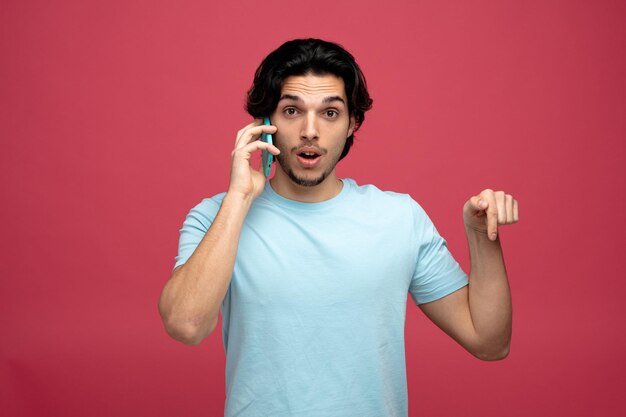 verraste jonge knappe man die naar de camera kijkt terwijl hij aan de telefoon praat en naar beneden wijst, geïsoleerd op rode achtergrond