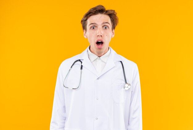 verraste jonge mannelijke arts die een medisch gewaad draagt met een stethoscoop geïsoleerd op een oranje muur
