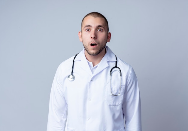 Verraste jonge mannelijke arts die medische mantel en stethoscoop om zijn hals draagt die voorzijde bekijkt die op witte muur wordt geïsoleerd