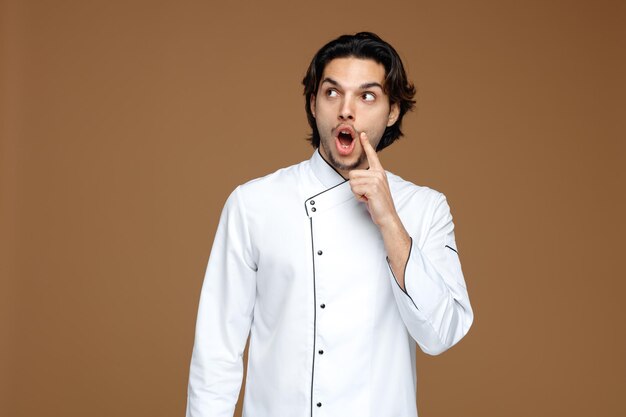 verraste jonge mannelijke chef-kok die uniform ontroerend gezicht draagt dat naar kant kijkt die op bruine achtergrond wordt geïsoleerd