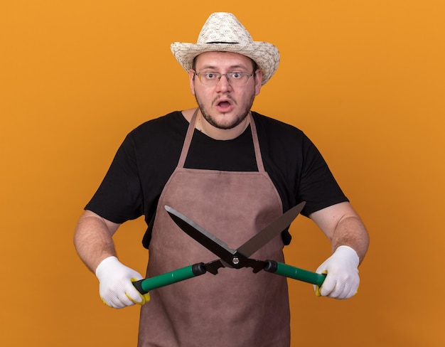 Gratis foto verraste jonge mannelijke tuinman die het tuinieren hoed en handschoenen draagt die tondeuse houden die op oranje muur wordt geïsoleerd