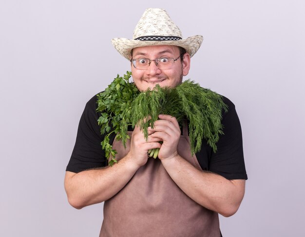 Verraste jonge mannelijke tuinman die tuinieren hoed draagt die dille met koriander rond gezicht houdt dat op witte muur wordt geïsoleerd