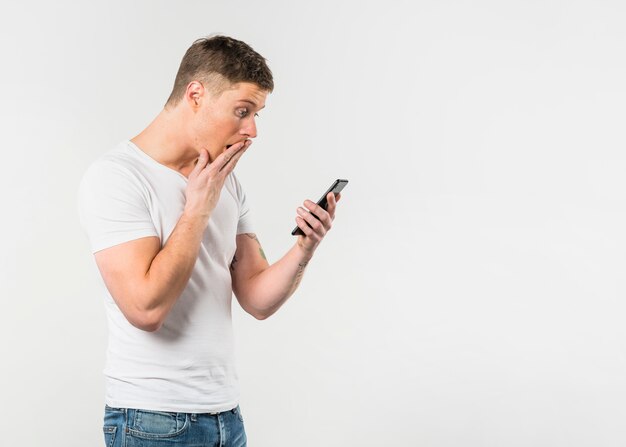 Verraste jonge mens die mobiele telefoon bekijkt die op witte achtergrond wordt geïsoleerd