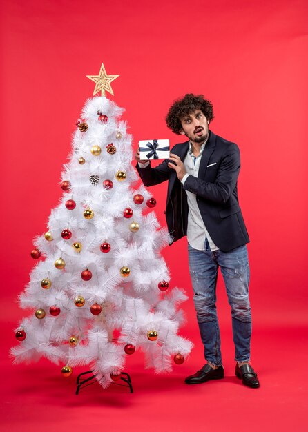 Verraste jonge mens die zijn gift vasthoudt en diep nadenkt staand dichtbij verfraaide witte kerstboom aan de rechterkant van rood