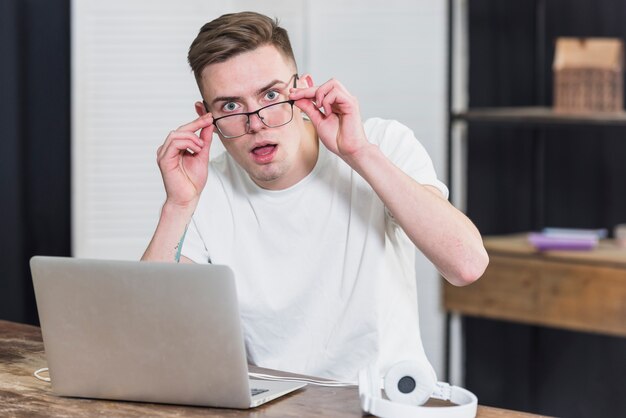 Verraste jonge mens met hoofdtelefoon en laptop op houten lijst die aan camera kijken
