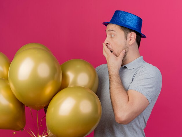 Verraste jonge partij kerel die blauwe hoed draagt ?? die en ballons houdt bedekt met hand kijkt die op roze wordt geïsoleerd