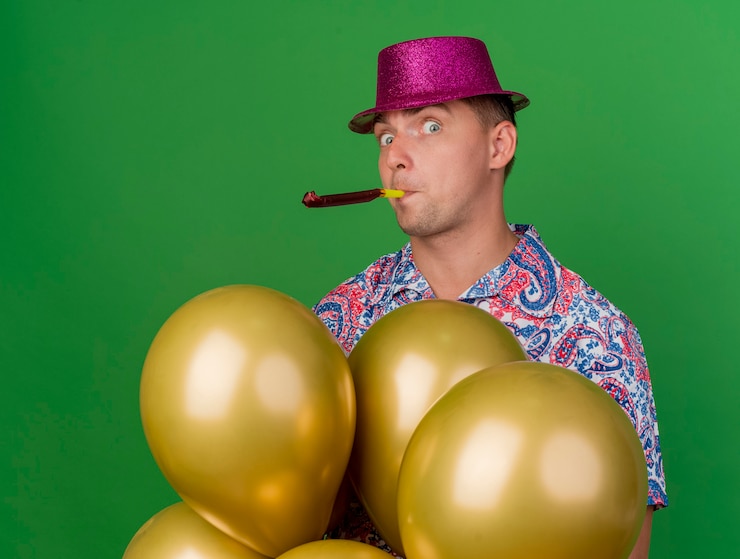 Gratis Foto verraste jonge partij kerel die roze hoed draagt die zich achter ballons bevindt en partijventilator blaast die op groen wordt geïsoleerd
