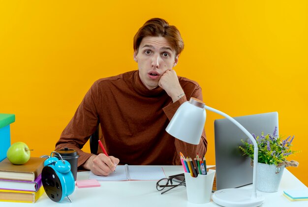 Verraste jonge studentenjongen zittend aan een bureau met schoolhulpmiddelen die iets op notitieboekje schrijven dat op gele muur wordt geïsoleerd