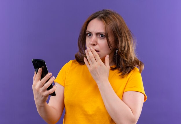 Verraste jonge toevallige vrouw die mobiele telefoon met hand op mond op geïsoleerde purpere muur houdt