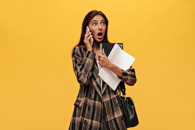 Verraste jonge vrouw in stijlvolle oversized jas praat over telefoon Geschokt meisje houdt notitieboekjes vast op gele achtergrond