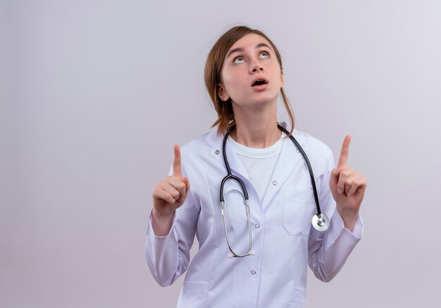 Verraste jonge vrouwelijke arts die medische mantel en stethoscoop draagt die omhoog kijkt en vingers op geïsoleerde witte muur met exemplaarruimte opheft