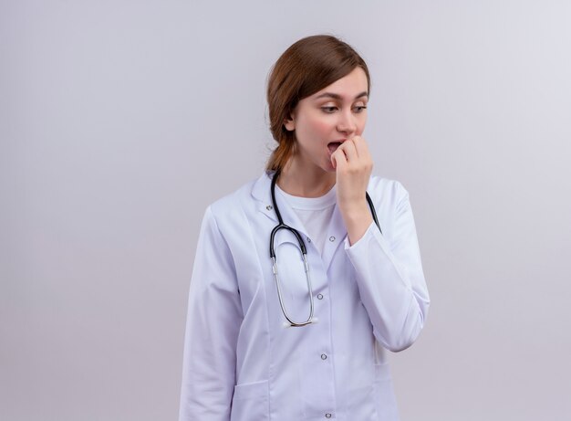 Verraste jonge vrouwelijke arts die medische mantel en stethoscoop draagt en hand op mond zet op geïsoleerde witte muur met exemplaarruimte