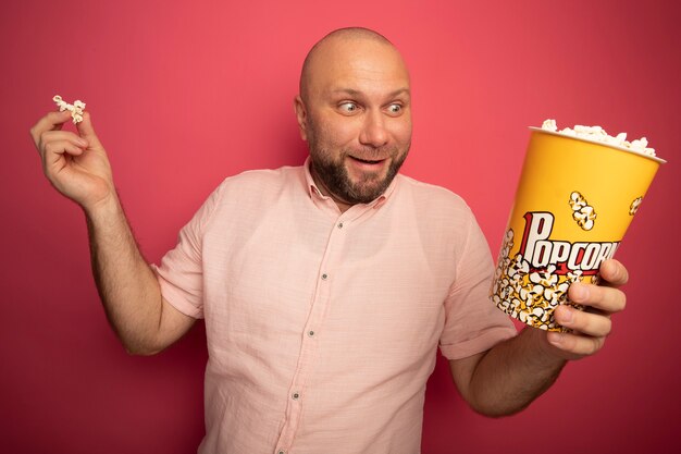 Verraste kale man van middelbare leeftijd die roze t-shirt draagt en emmer popcorn met popcornvrede houdt die op roze muur wordt geïsoleerd