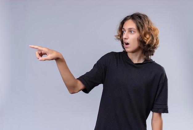 Verraste kerel met lang haar in zwart t-shirt wijst naar kant op witte achtergrond