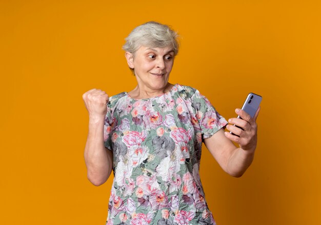 Verraste oudere vrouw heft vuist houden en kijken naar telefoon geïsoleerd op oranje muur