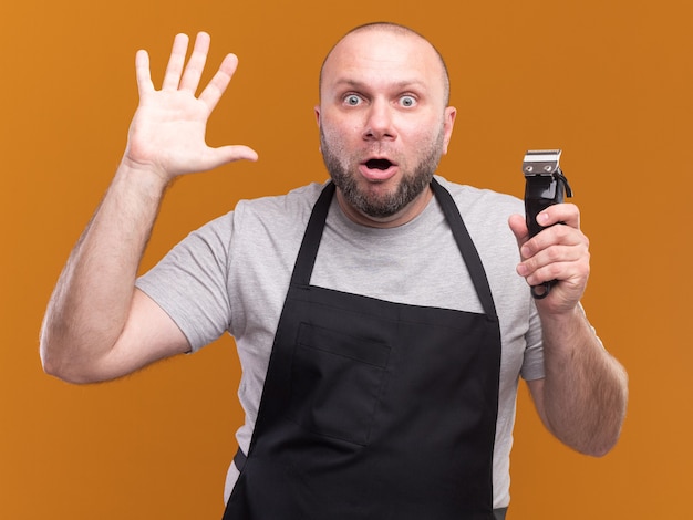 Verraste Slavische mannelijke kapper van middelbare leeftijd in eenvormige clipper van het holdingshaar die vijf toont die op oranje muur worden geïsoleerd