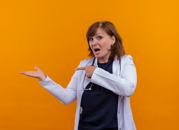 Verraste vrouw arts die op middelbare leeftijd medische mantel en stethoscoop draagt die met hand en vinger aan linkerkant op geïsoleerde oranje muur richt