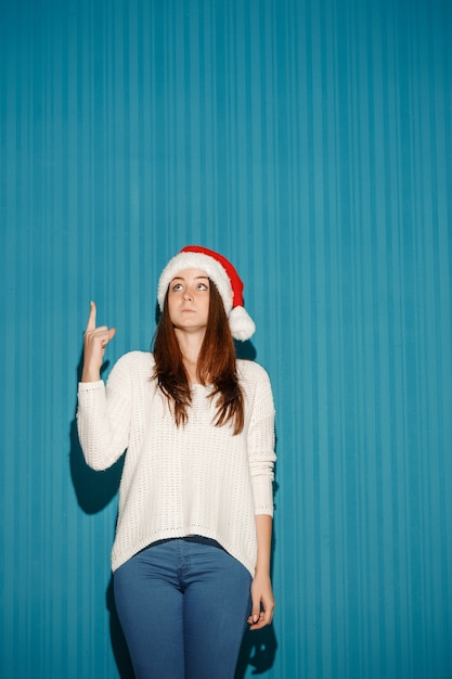 Gratis foto verraste vrouw die een kerstmuts draagt die op blauwe studioachtergrond wijst