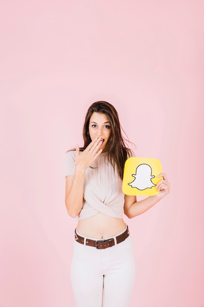 Gratis foto verraste vrouw die snapchat-pictogram op roze achtergrond houdt