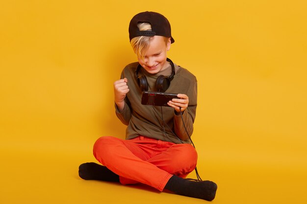 verrukt jongetje kleedt oranje broek en groen shirt spelen van videogame op mobiel en balde vuist zittend op de vloer met gekruiste benen geïsoleerd op geel