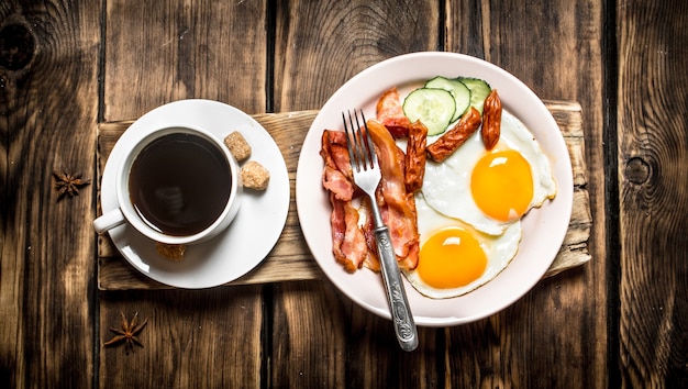 Vers ontbijt. kopje koffie, gebakken spek met eieren en rookworst. op houten achtergrond.