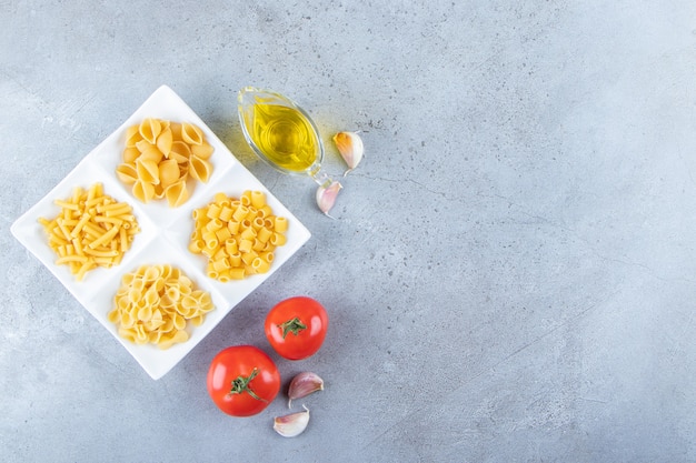 Verschillende soorten rauwe droge pasta met verse rode tomaten en olie op een stenen ondergrond.