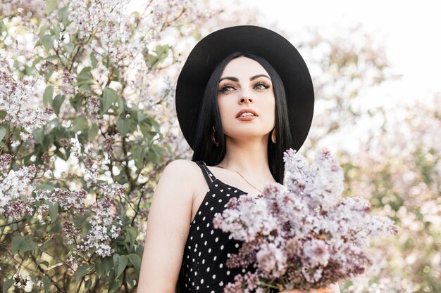 Verse portret aantrekkelijke vrouw in zwarte trendy jurk in hoed met mooie bloemen in handen op achtergrond van lila verbazingwekkende bloeiende bomen in park bij zonsondergang. mooi meisje in tuin op heldere zonnige dag