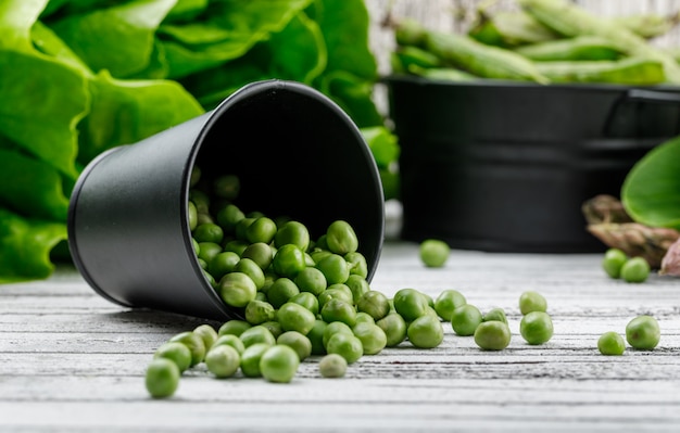 Gratis foto verspreide erwten uit een emmer met asperges, paksoi, sla, groene peulen zijaanzicht op een houten muur