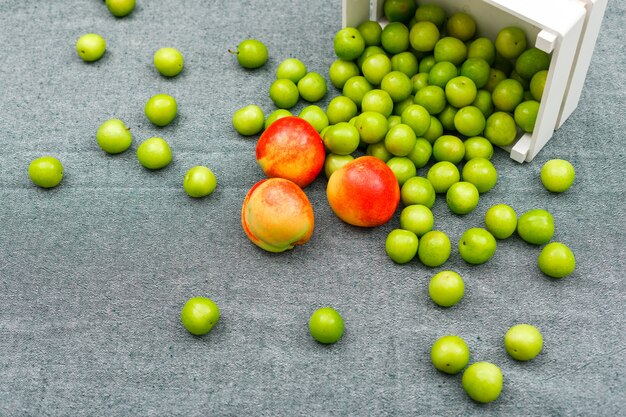 Verspreide groene pruimen met smakelijke perziken in een witte rechthoekkom op grijze, hoge hoekmening.