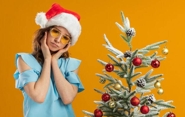 Verstoorde jonge vrouw in blauwe top en kerstmuts met een gele bril met een droevige uitdrukking die naast een kerstboom over een oranje muur staat
