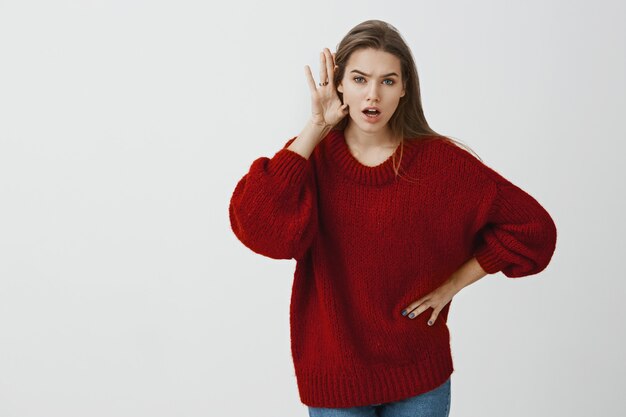 Vertel me alles wat hij tegen je zei. Binnenopname van geschokte en intens charmante vrouwelijke collega in losse rode trui, buigend naar camera, handpalm bij oor houdend en geruchten of roddels verspreiden