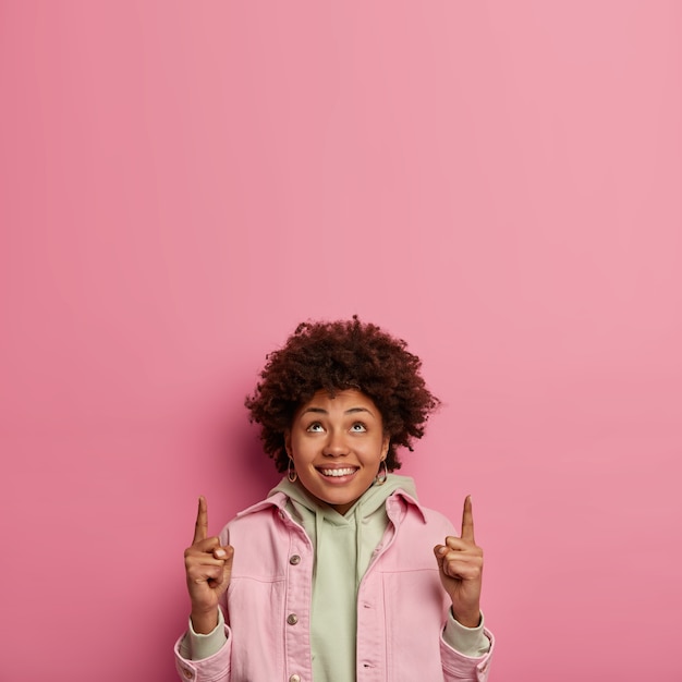 Verticaal beeld van lachende optimistische gekrulde vrouw wijst beide wijsvingers naar boven, heeft een vrolijke uitdrukking, adverteert cool promo naar boven