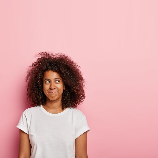 Verticaal beeld van vrij dromerige jonge vrouw houdt lippen gedrukt, boven gefocust met ogen vol geluk, stelt zich iets aangenaams voor, heeft volumineus donker haar, draagt een casual wit t-shirt.