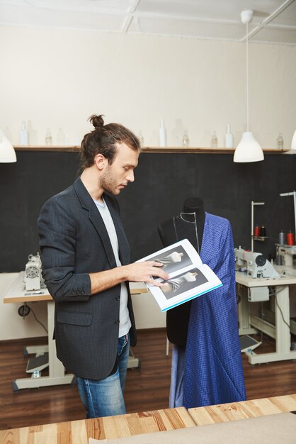 Verticaal portret van jonge succesvolle professionele mannelijke kleermaker die aan blauwe kleding voor klant werkt, herinnerend sommige naaitechnieken door te kijken door leerboek.