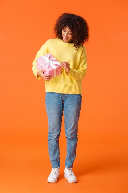 Verticaal schot over de volledige lengte schattig en gelukkig feestvarken uitpakken van cadeau, lachende Afro-Amerikaanse vrouw met krullend haar in trui, roze aanwezig nieuwsgierig wat erin zit, oranje muur.