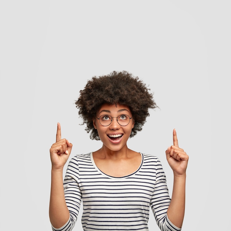 Gratis Foto verticaal schot van aangenaam ogende afro-amerikaanse vrouw met vrolijke uitdrukking, heeft krullend haar, wijst naar boven met wijsvingers
