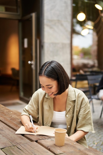 Verticaal schot van een aziatische studente die iets in een notitieboekje schrijft en aantekeningen maakt terwijl ze glimla