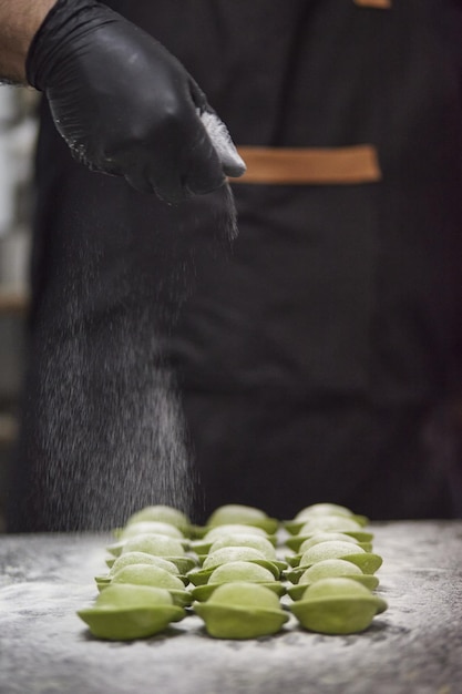 Verticaal schot van een mannenhand in een zwarte handschoen die rauwe groene cappelletti licht bestrooit met bloem