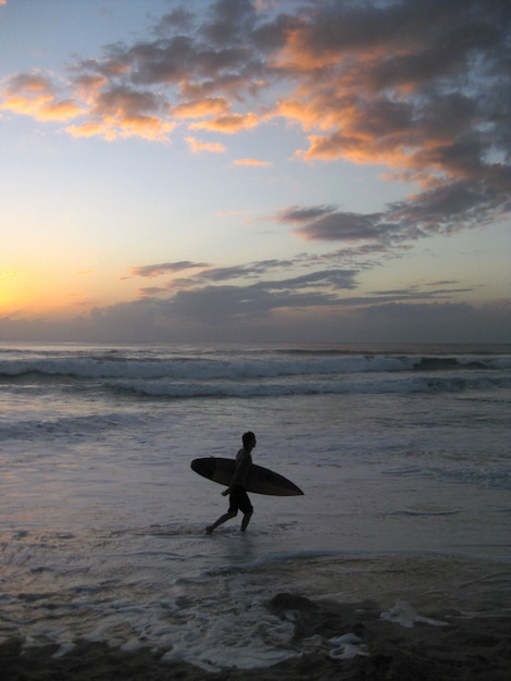 Verticaal schot van een persoon die een surfplank houdt lopend dichtbij een golvende overzees tijdens zonsondergang