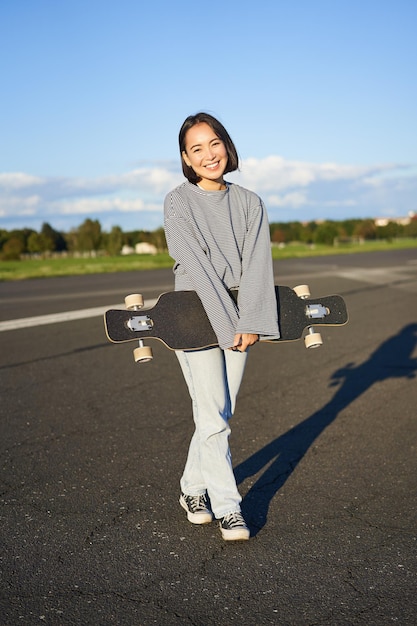 Gratis foto verticaal schot van een skatermeisje dat poseert met een longboard dat op een lege weg in de buitenwijken lacht en aziatisch glimlacht