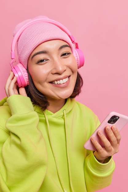 Verticaal schot van gelukkige jonge aziatische vrouw gekleed in vrijetijdskleding geniet van favoriete muziek draagt koptelefoon op oren met goede geluidskwaliteit houdt moderne smartphone geïsoleerd over roze muur.