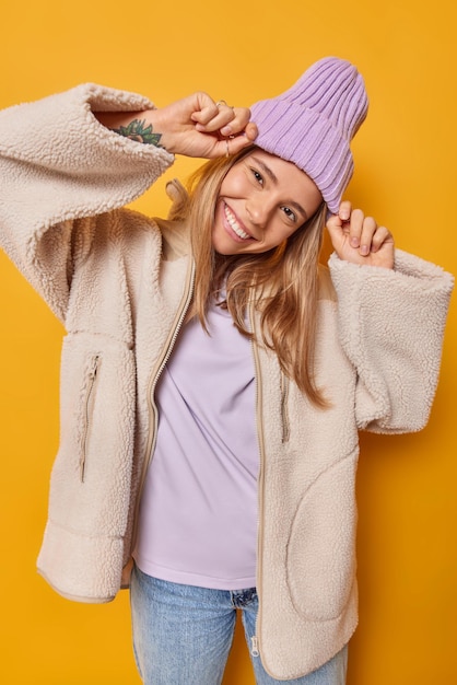Verticaal schot van gelukkige jonge europese vrouw houdt handen op hoed kantelt hoofd en glimlacht graag warme bontjas geïsoleerd over gele achtergrond geniet van goede dag. mensen en kleding concept