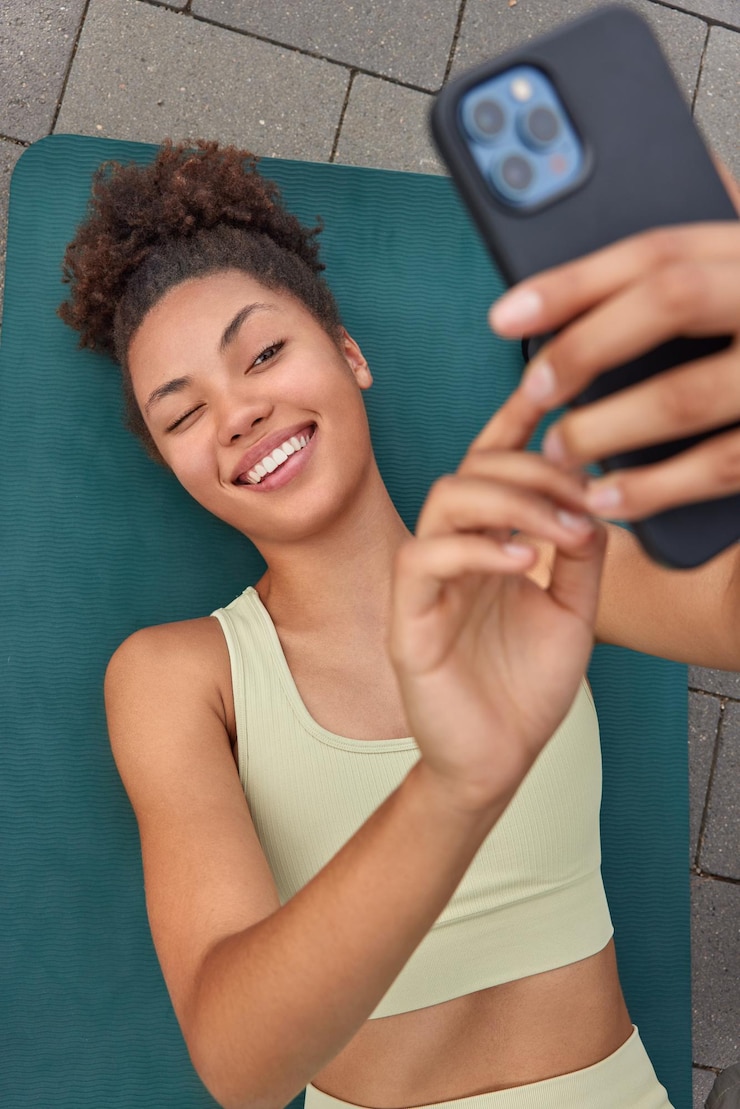Gratis Foto verticaal schot van gelukkige sportieve vrouw knipoogt naar smartphonecamera terwijl selfie rust neemt na training met gespierde armen gekleed in activewear ligt op fitnessmat. sport- en technologieconcept