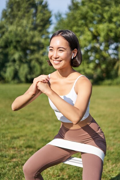 Verticaal schot van jonge fitte vrouw doet squats in het park met behulp van een rekbare band op benen die tevreden glimlachen
