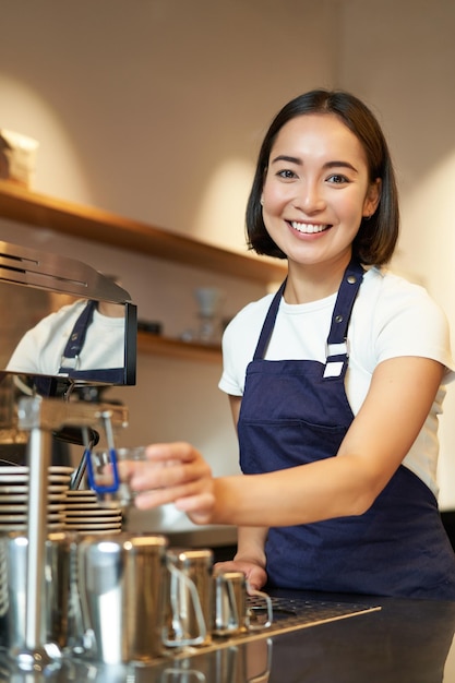 Verticaal schot van jonge professionele glimlachende meisjesbarista die in koffie dampende melk voor cappuccino werkt