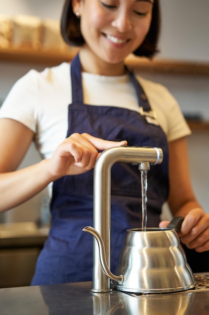 Verticaal schot van meisjesbarista in café die water uit de kraan giet met behulp van een waterkoker om filterkoffie te zetten