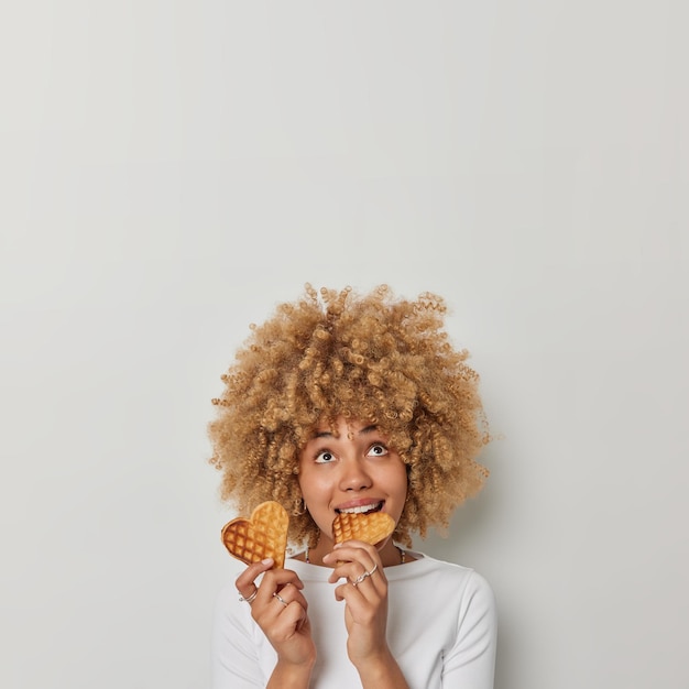 Gratis foto verticaal schot van mooie vrouw met krullend haar bijt smakelijke hartvormige wafels boven gericht geniet van het eten van heerlijk eten gekleed in casual trui geïsoleerd op witte achtergrond lege ruimte